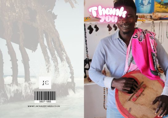 A man holds a traditional musical instrument, wearing a pink scarf, near a shop interior.